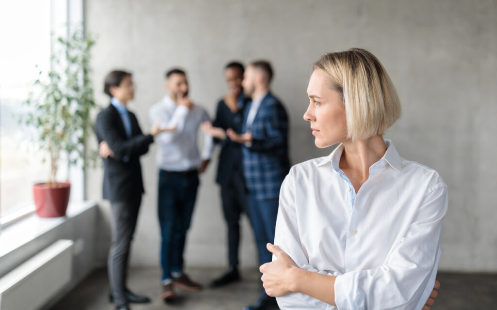 Male Coworkers Whispering Behind Back Of Unhappy Businesswoman Spreading Rumors And Gossips Standing In Modern Office